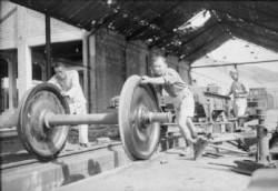 Leading Aircraftman L Moulden, a former blacksmith, helps roll a newly repaired bogey unit for a carriage or flatbed truck into place at the Kowloon Railway's repair sheds, Hong Kong. This railway was immobilised during the Japanese occupation, services were restored under Royal Air Force supervision after the liberation.Made by: Royal Air Force official photographer